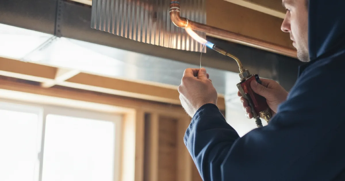 A plumber soldering a repair coupling into a copper supply line in an open basement ceiling, heat shield in place behind the joint, clean workmanship